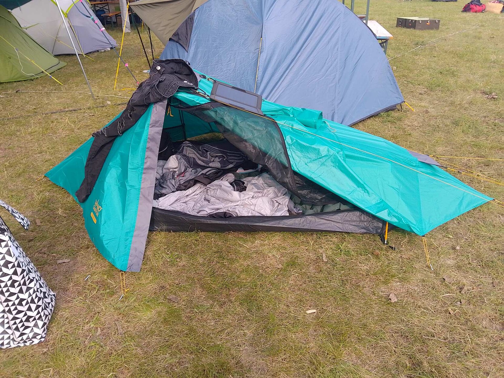 kleines türkises Zelt auf dem eine Hose zum lüften liegt und eine Solazeller liegt. Vorne ist das Zelt ofen und es ist ein Schlafsack zu sehen.
